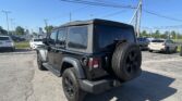 Rear three-quarter view of a black Jeep Wrangler with a soft top in a parking lot, spare tire on the back, blue sky above.