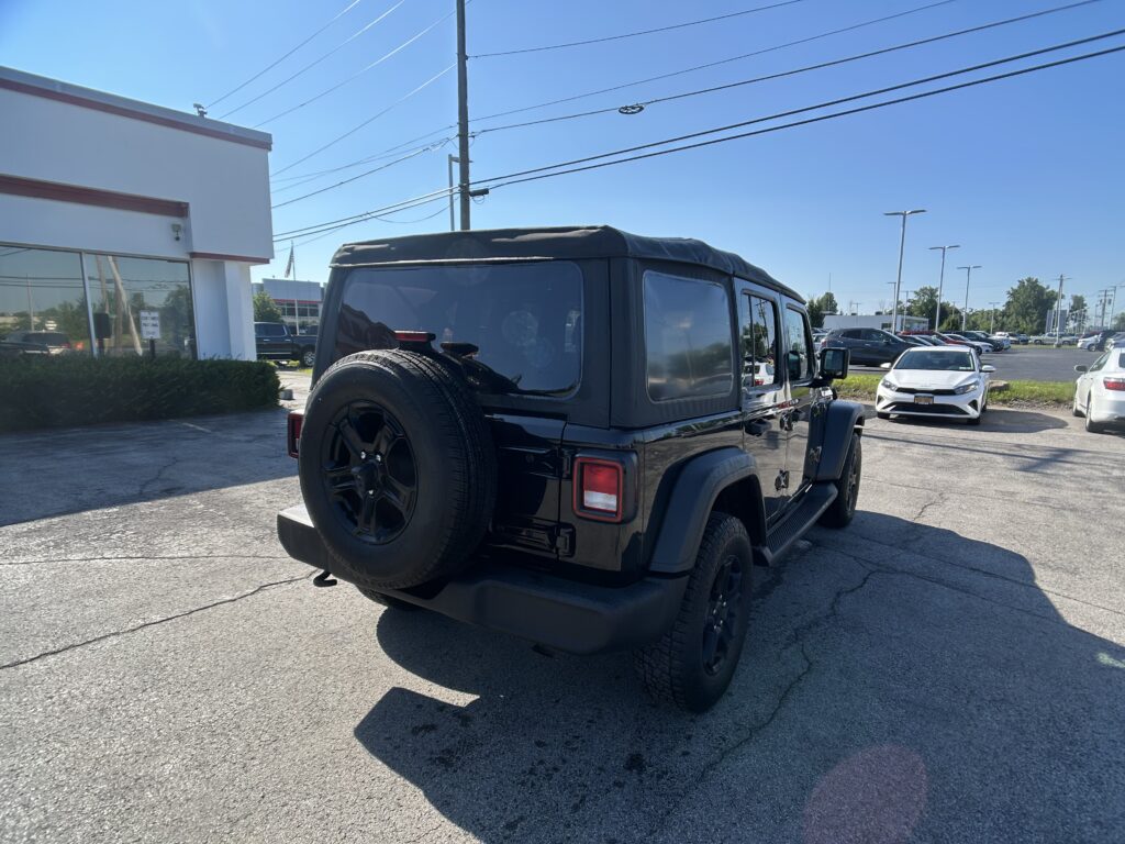 Black Jeep Wrangler with spare tire mounted on the back, parked in a dealership lot beside a white building under a clear blue sky.