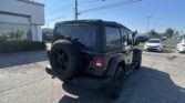 Black Jeep Wrangler with spare tire mounted on the back, parked in a dealership lot beside a white building under a clear blue sky.