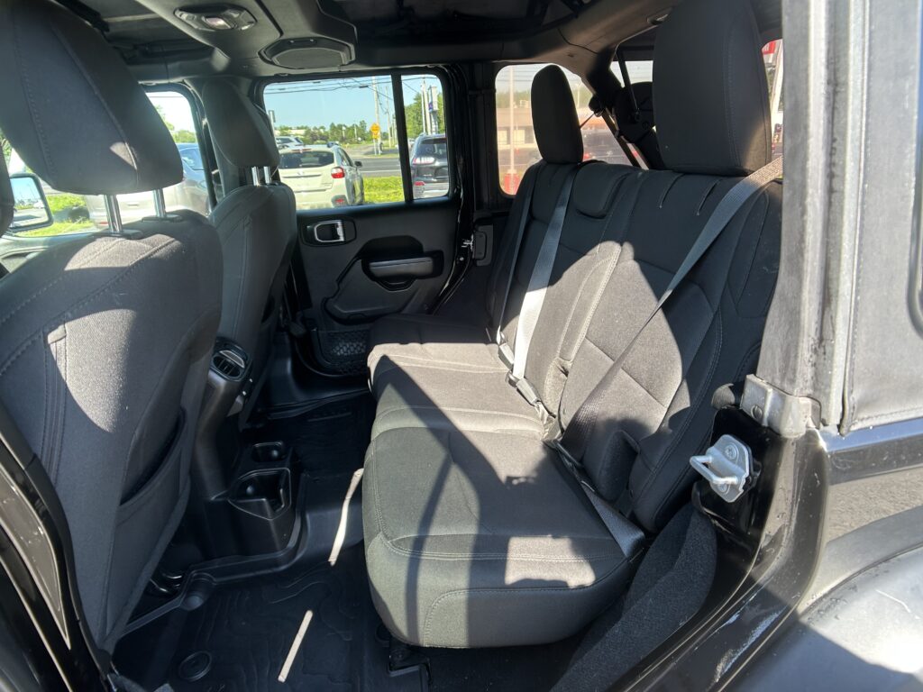 Back seat area of a black SUV with three head restraints and visible seat belts, viewed from the open rear door; outside cars in a parking lot visible through windows.
