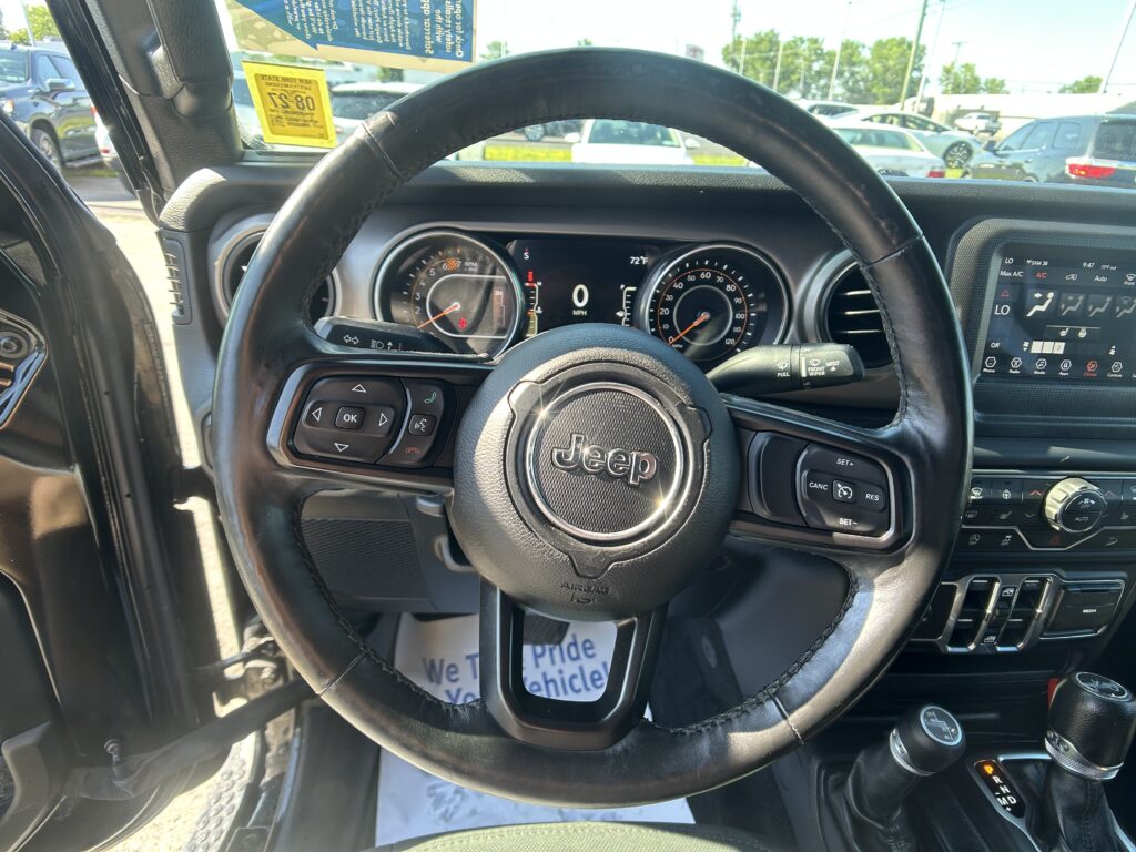 Interior view of a Jeep SUV steering wheel and dashboard, showing the instrument cluster, central touchscreen, and gear selector with PRND, while parked cars are visible through the windshield.