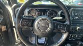 Interior view of a Jeep SUV steering wheel and dashboard, showing the instrument cluster, central touchscreen, and gear selector with PRND, while parked cars are visible through the windshield.
