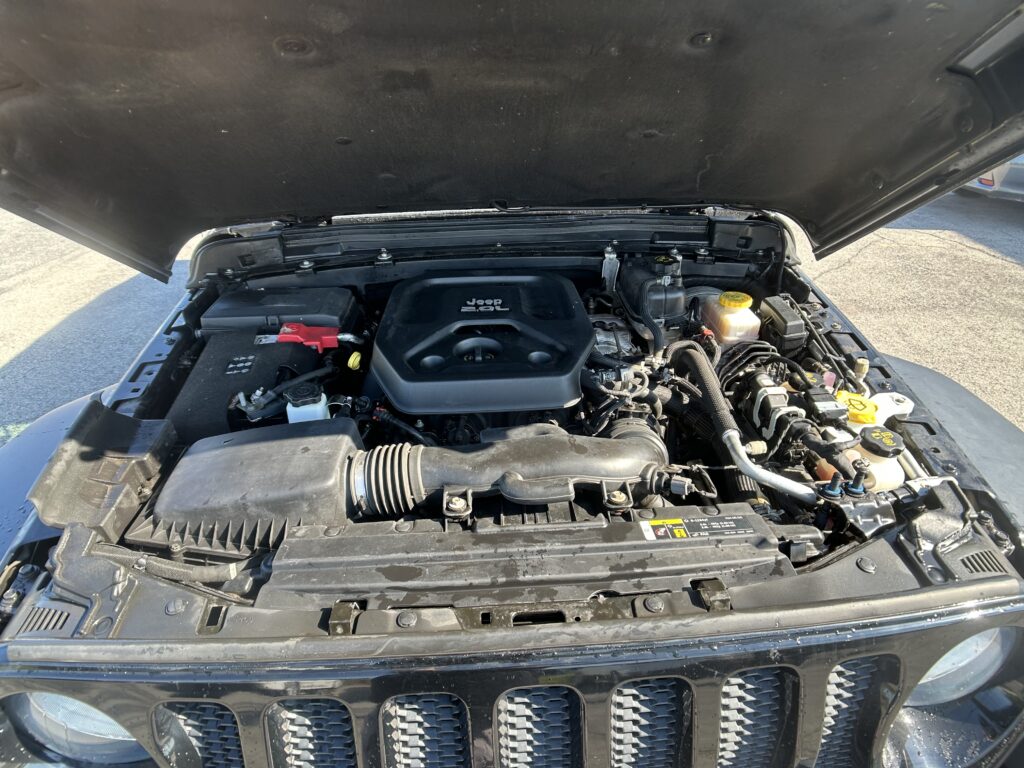 Under-hood view of a Jeep with the hood raised, showing the engine bay, air intake, battery, and fluid reservoirs visible under the engine cover.