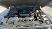 Under-hood view of a Jeep with the hood raised, showing the engine bay, air intake, battery, and fluid reservoirs visible under the engine cover.