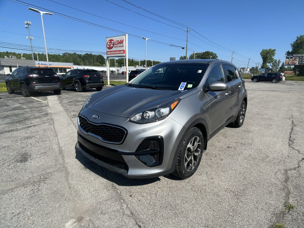 Front-right view of a silver Kia SUV parked in a dealership lot with other cars and an EZ Loan Auto Sales sign in the background on a sunny day.