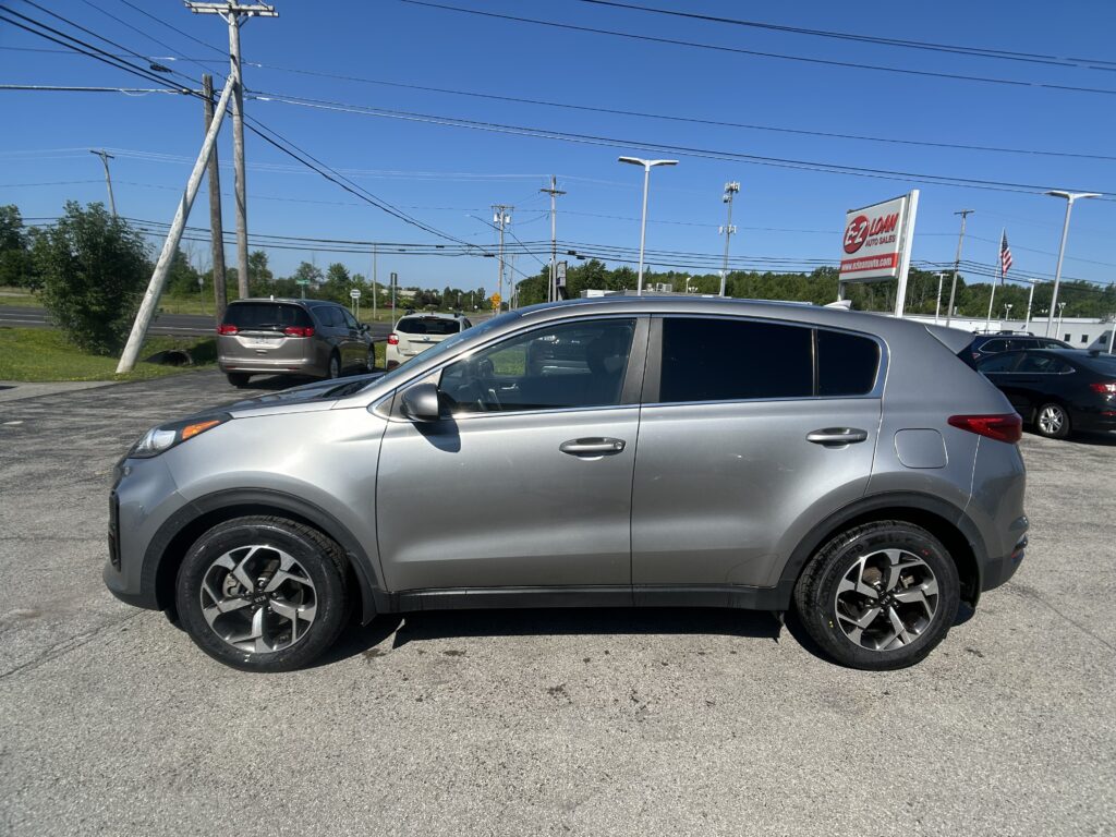 Silver SUV in the foreground at a car dealership lot, with other vehicles, utility poles, and a blue sky in the background, and an EZ LOAN Auto Sales sign visible in the distance.