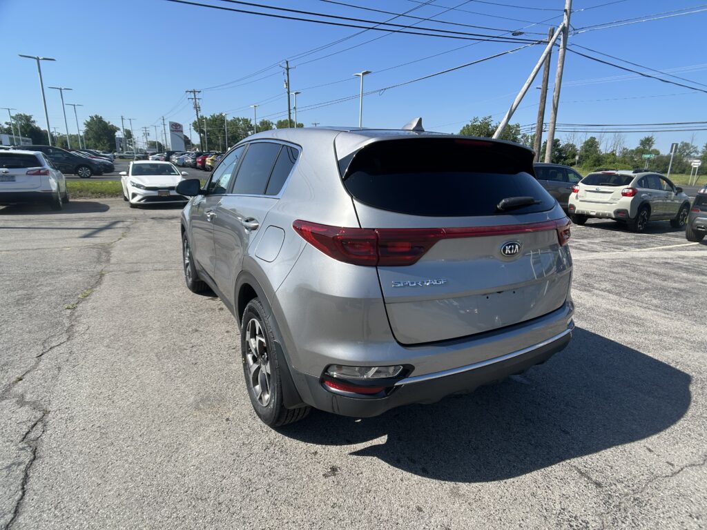 Silver Kia Sportage SUV parked at a car dealership lot, rear view in foreground with other cars and power lines in the background.