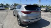 Silver Kia Sportage SUV parked at a car dealership lot, rear view in foreground with other cars and power lines in the background.