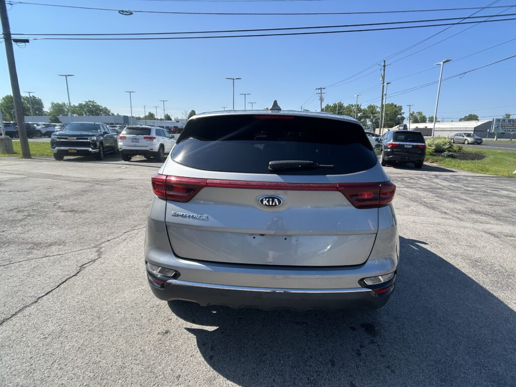 Rear view of a silver Kia Sportage parked in a car dealership lot, with other cars and power lines overhead under a clear blue sky.