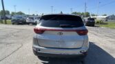 Rear view of a silver Kia Sportage parked in a car dealership lot, with other cars and power lines overhead under a clear blue sky.