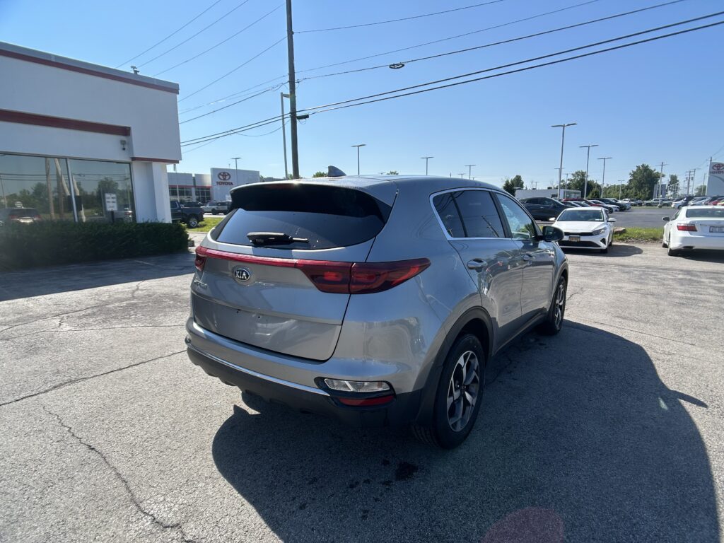 Rear three-quarter view of a silver Kia SUV parked in a dealership lot, with a Toyota building and signage in the background under a clear blue sky.