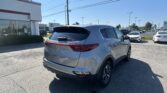 Rear three-quarter view of a silver Kia SUV parked in a dealership lot, with a Toyota building and signage in the background under a clear blue sky.