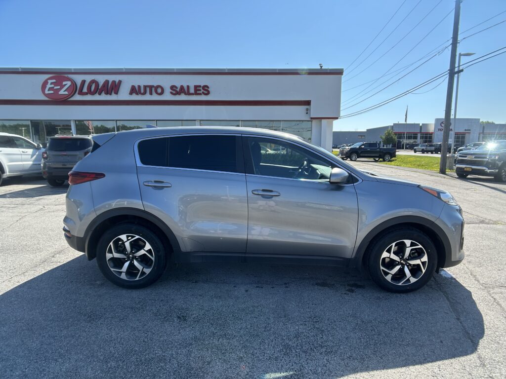 Silver SUV parked in the foreground outside an auto dealership with an 'E-Z LOAN AUTO SALES' sign on the building under a clear blue sky.