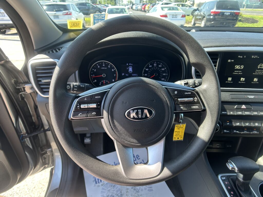 View from the driver's seat showing a Kia steering wheel, instrument cluster, and dashboard with a parked-car lot visible through the front windshield on a sunny day
