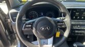 View from the driver's seat showing a Kia steering wheel, instrument cluster, and dashboard with a parked-car lot visible through the front windshield on a sunny day