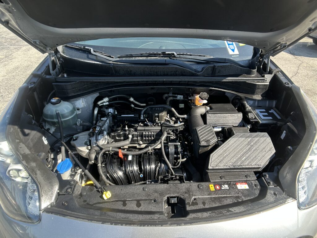 Car engine bay with the hood raised, showing the engine, hoses, battery, and coolant reservoir visible in the front compartment.