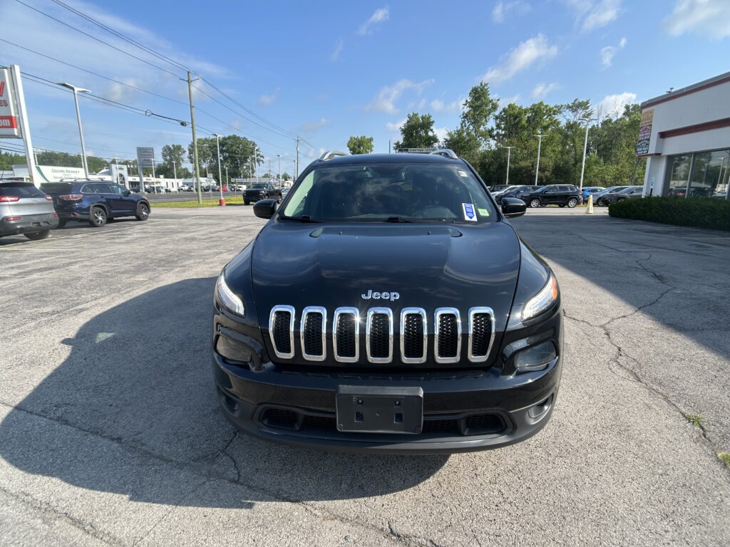 Front view of a black Jeep SUV (Grand Cherokee) parked in a dealership lot on a sunny day with other cars and a building in the background.