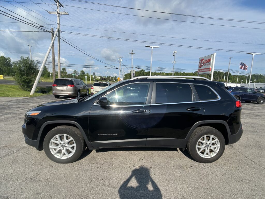 Black Jeep Cherokee in a car dealership lot, with other vehicles, power lines, and an Auto Sales sign in the background. American flag nearby.