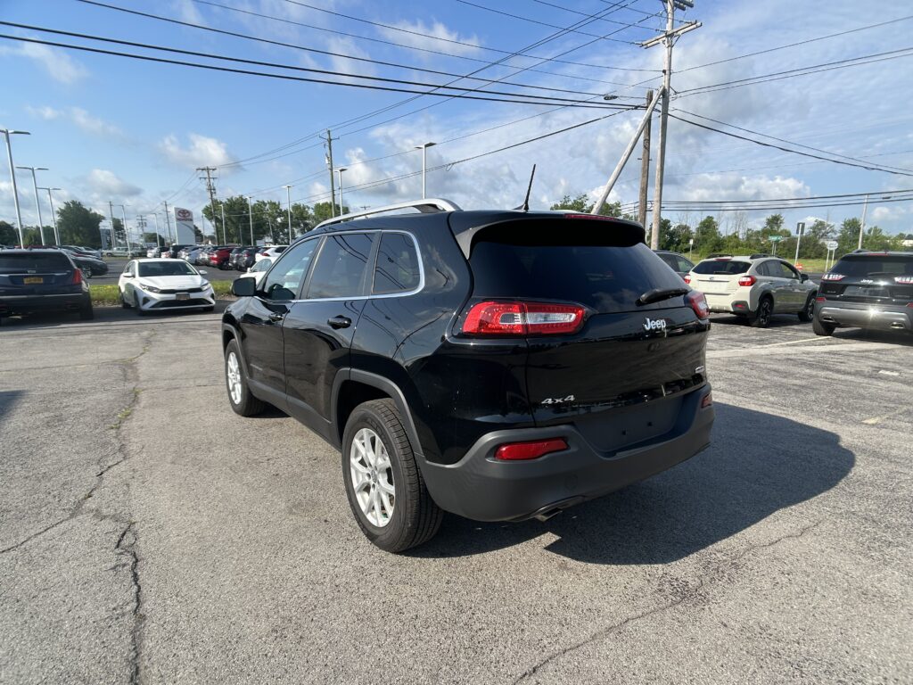 Black Jeep SUV in the foreground of a car dealership lot with other cars and power lines overhead under a blue sky.