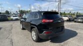 Black Jeep SUV in the foreground of a car dealership lot with other cars and power lines overhead under a blue sky.