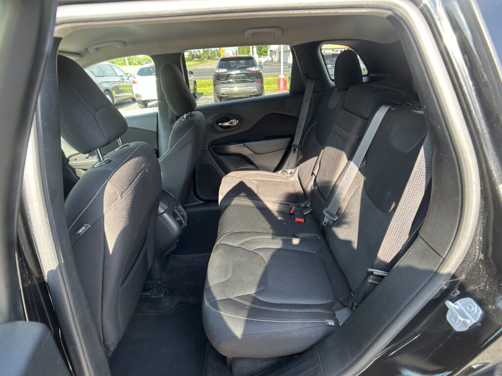 Rear passenger area of a black SUV with dark fabric seats and visible seat belts, looking toward the back seats from the open rear door; outside cars and a street are visible through the windows.