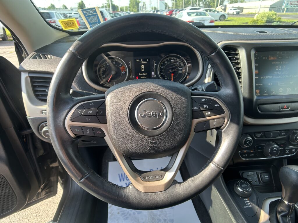 Driver's perspective inside a Jeep showing the steering wheel, instrument cluster, and right-side infotainment screen; outside parking lot visible through the windshield.