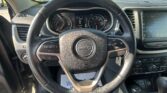 Driver's perspective inside a Jeep showing the steering wheel, instrument cluster, and right-side infotainment screen; outside parking lot visible through the windshield.