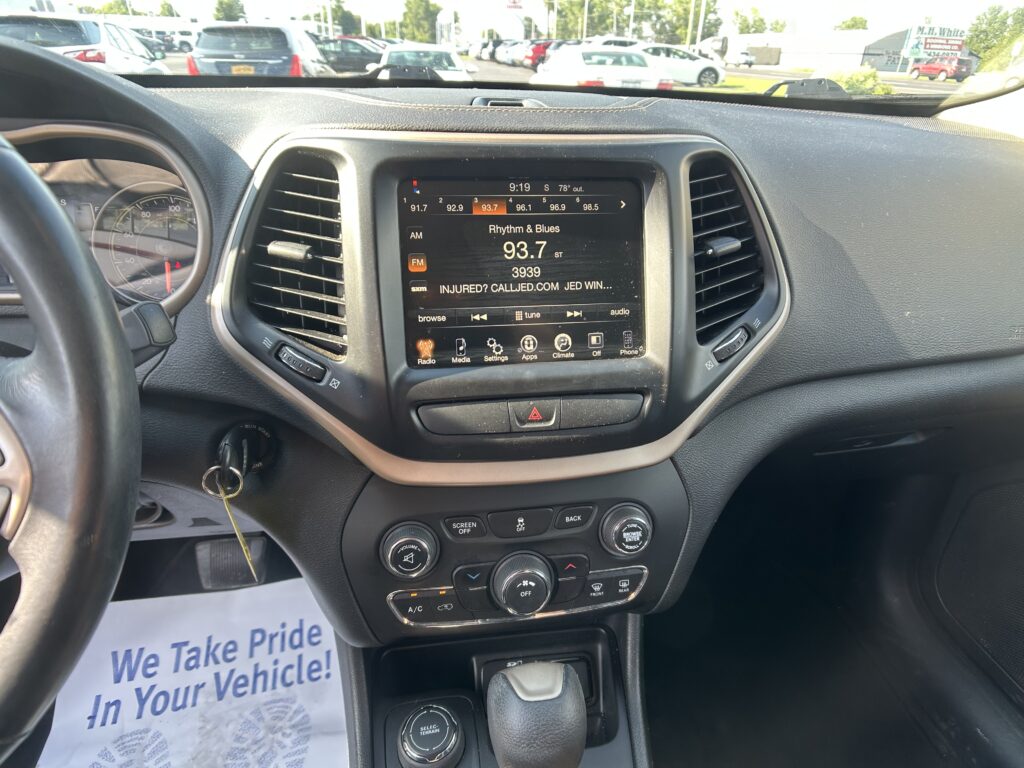 Inside a car cockpit with the center touchscreen displaying FM radio 93.7 Rhythm & Blues and a parking lot visible through the windshield