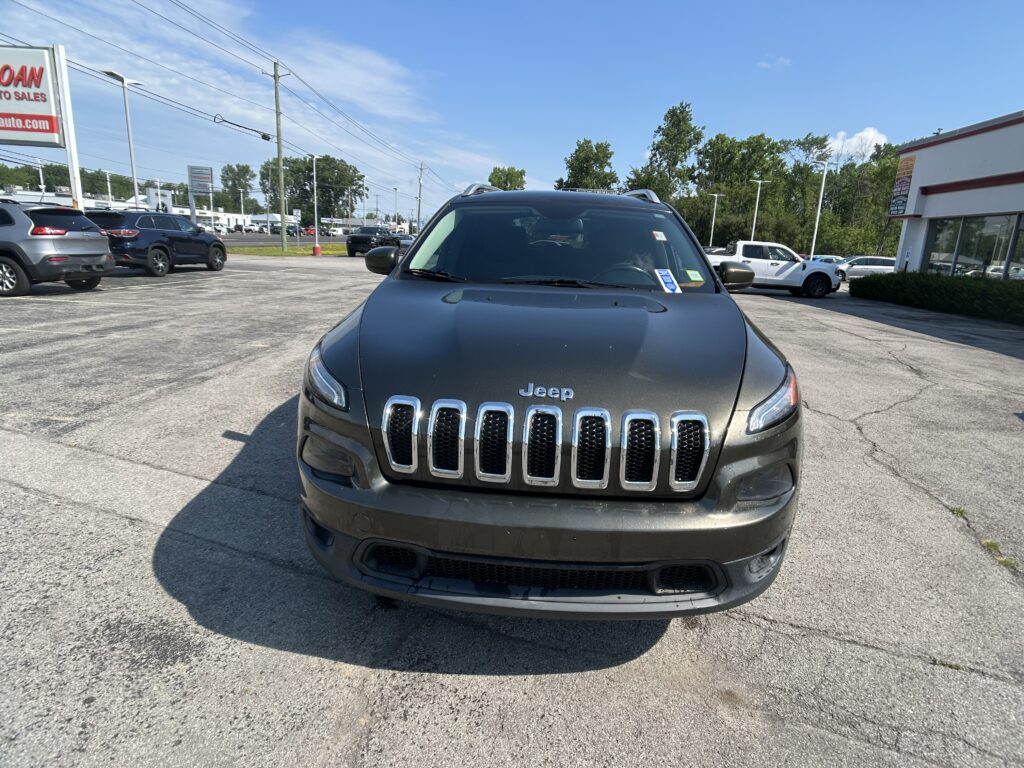 Front view of a dark gray Jeep SUV parked in a car dealership lot with other vehicles and a building in the background on a sunny day.