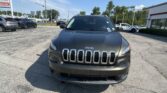 Front view of a dark gray Jeep SUV parked in a car dealership lot with other vehicles and a building in the background on a sunny day.