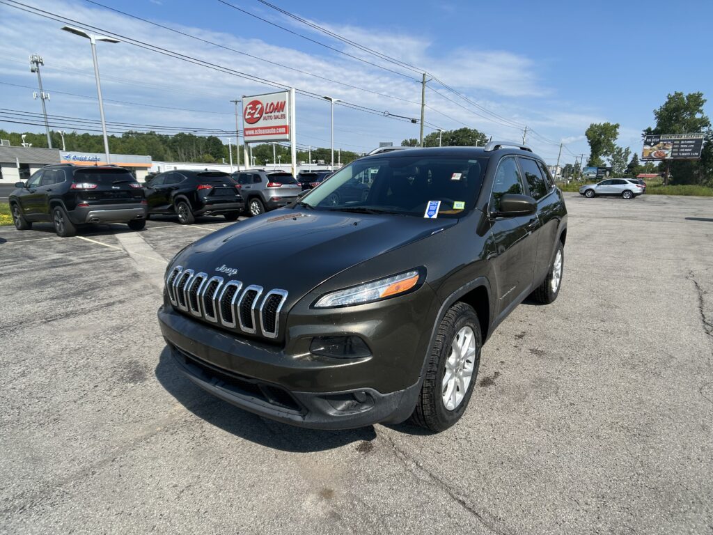 Close-up view of a dark Jeep SUV in a car lot with other vehicles and an EZ LOAN Auto Sales sign in the background under a blue sky.