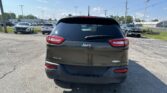 Rear view of a dark gray Jeep SUV in a parking lot, other vehicles and dealership buildings visible in the background.