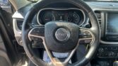 Driver's view of the Jeep steering wheel and dashboard in a car parked in a lot, with a row of cars visible through the windshield.