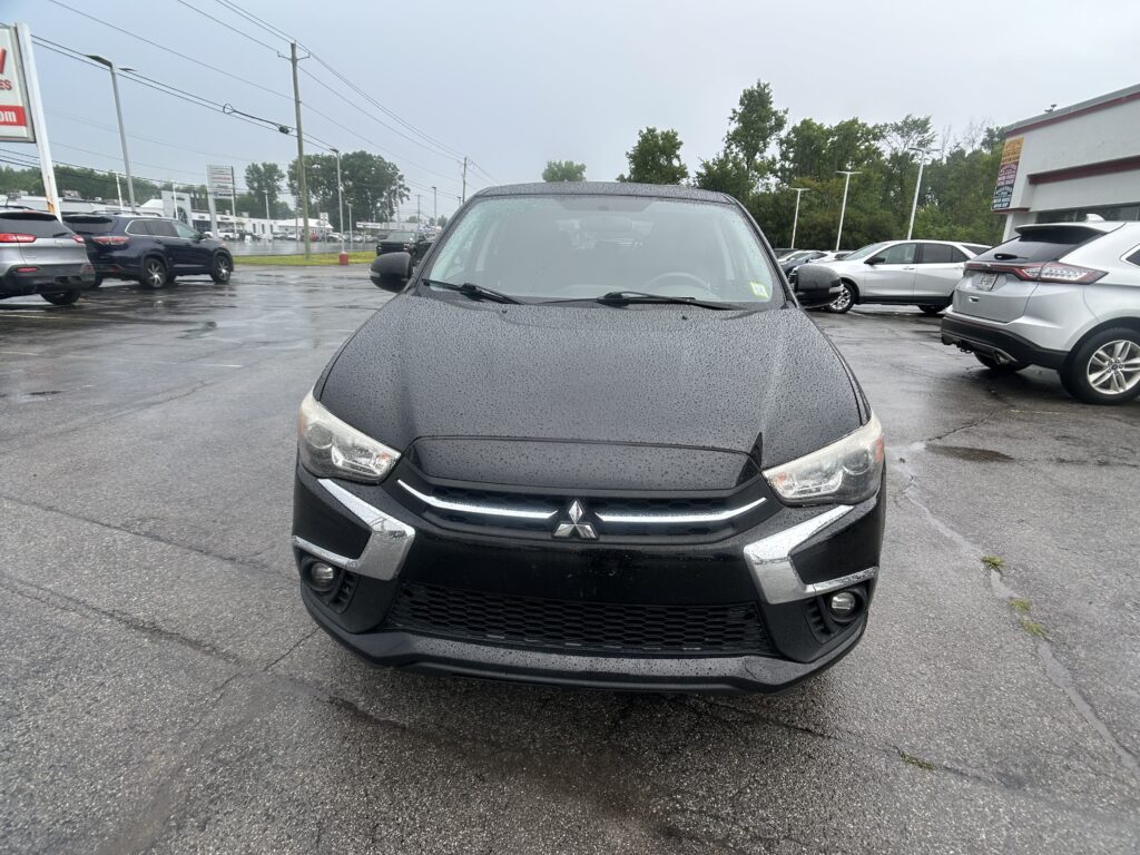 Front view of a black Mitsubishi SUV parked on a wet dealership lot with other cars and a building in the background, overcast sky above.