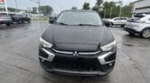 Front view of a black Mitsubishi SUV parked on a wet dealership lot with other cars and a building in the background, overcast sky above.