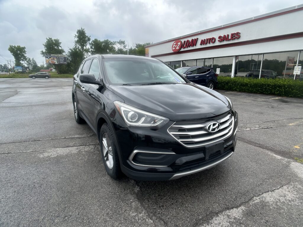 Gray Hyundai SUV in the foreground is parked at a car dealership with an E-Z Loan Auto Sales building behind it on a cloudy day.