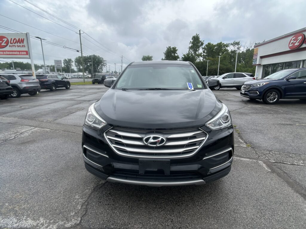 Front view of a black Hyundai SUV in a dealership lot with other cars and a sign in the background under cloudy skies, wide-angle view.