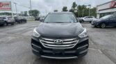 Front view of a black Hyundai SUV in a dealership lot with other cars and a sign in the background under cloudy skies, wide-angle view.