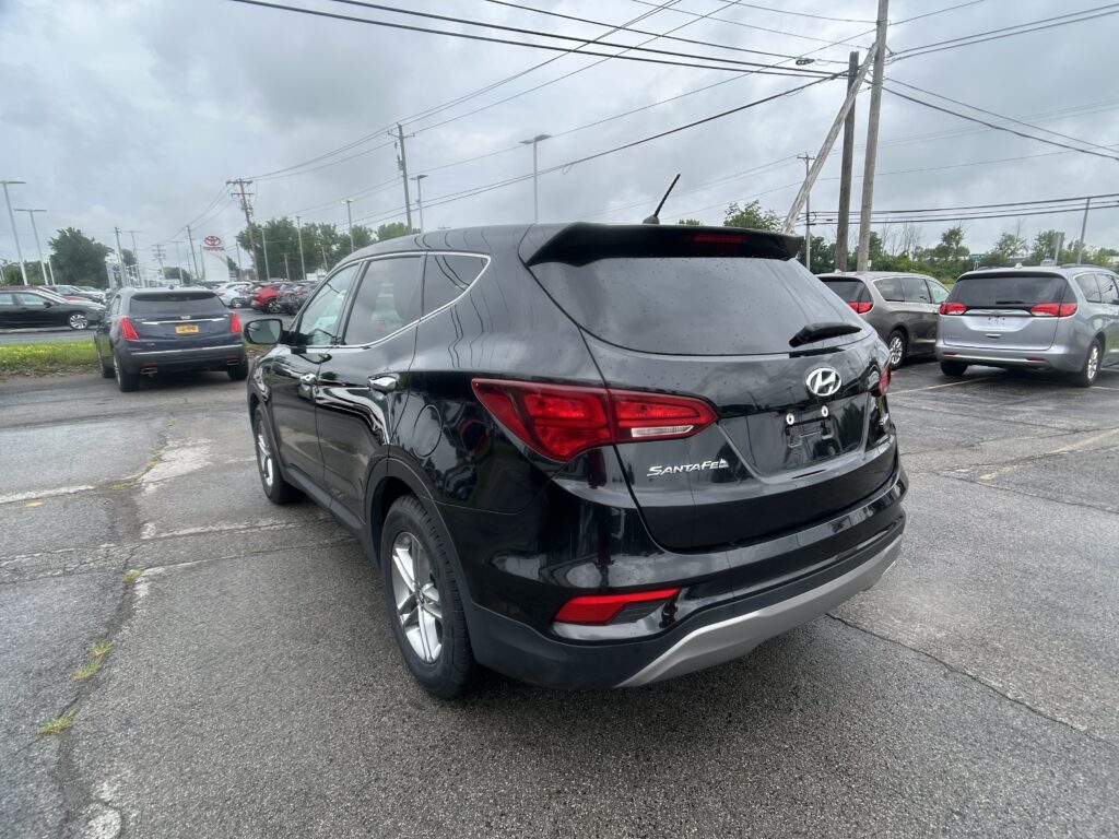 Rear view of a black Hyundai Santa Fe SUV in a busy dealership lot on a cloudy day, with other cars nearby and power lines above.