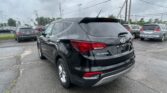 Rear view of a black Hyundai Santa Fe SUV in a busy dealership lot on a cloudy day, with other cars nearby and power lines above.