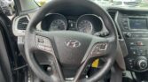 Close-up of a car steering wheel with the Hyundai emblem, showing the instrument cluster and center console in a showroom/lot setting outside the windshield.