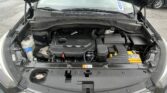 Front view with hood up showing the car's engine bay: battery on the right, coolant reservoir on the left, and various hoses and components visible under the hood with windshield and wipers in the background.