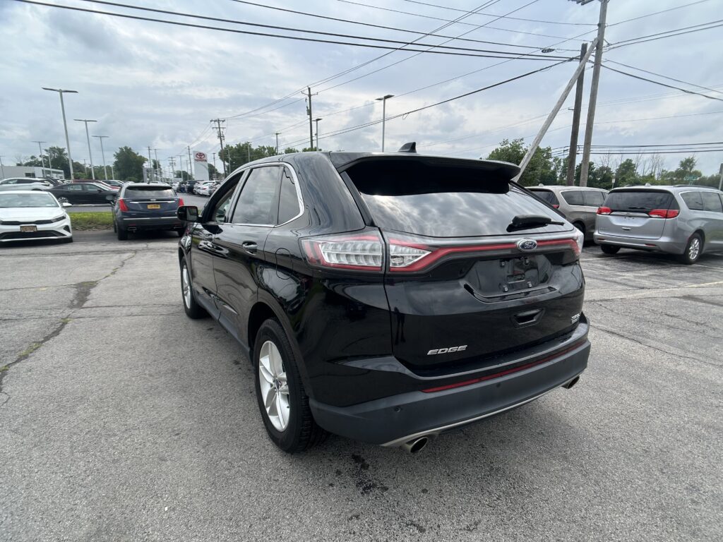 Rear view of a black Ford Edge SUV in a crowded car lot under cloudy skies, with other vehicles and power lines in the background.