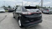 Rear view of a black Ford Edge SUV in a crowded car lot under cloudy skies, with other vehicles and power lines in the background.
