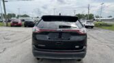 Rear view of a black Ford Edge SUV in a car dealership parking lot, with other cars and overhead power lines.