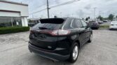 Rear view of a black Ford Edge SUV in a dealership parking lot with other cars and a white building to the left.