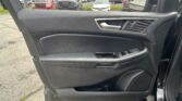 Inside a car door panel with a silver handle, window controls, and a side mirror; parking lot with cars visible outside the window.