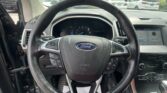 Driver's-seat view of a Ford steering wheel with dashboard, controls, and infotainment screen visible, car lot outside through the windshield.