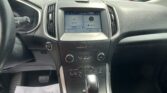 Interior of a car cockpit with a center touchscreen display and climate controls, view of a busy parking lot through the windshield