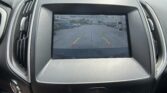 Rearview camera display on car screen showing parking guidelines in a lot, with 'Please check surroundings for safety' text at top.